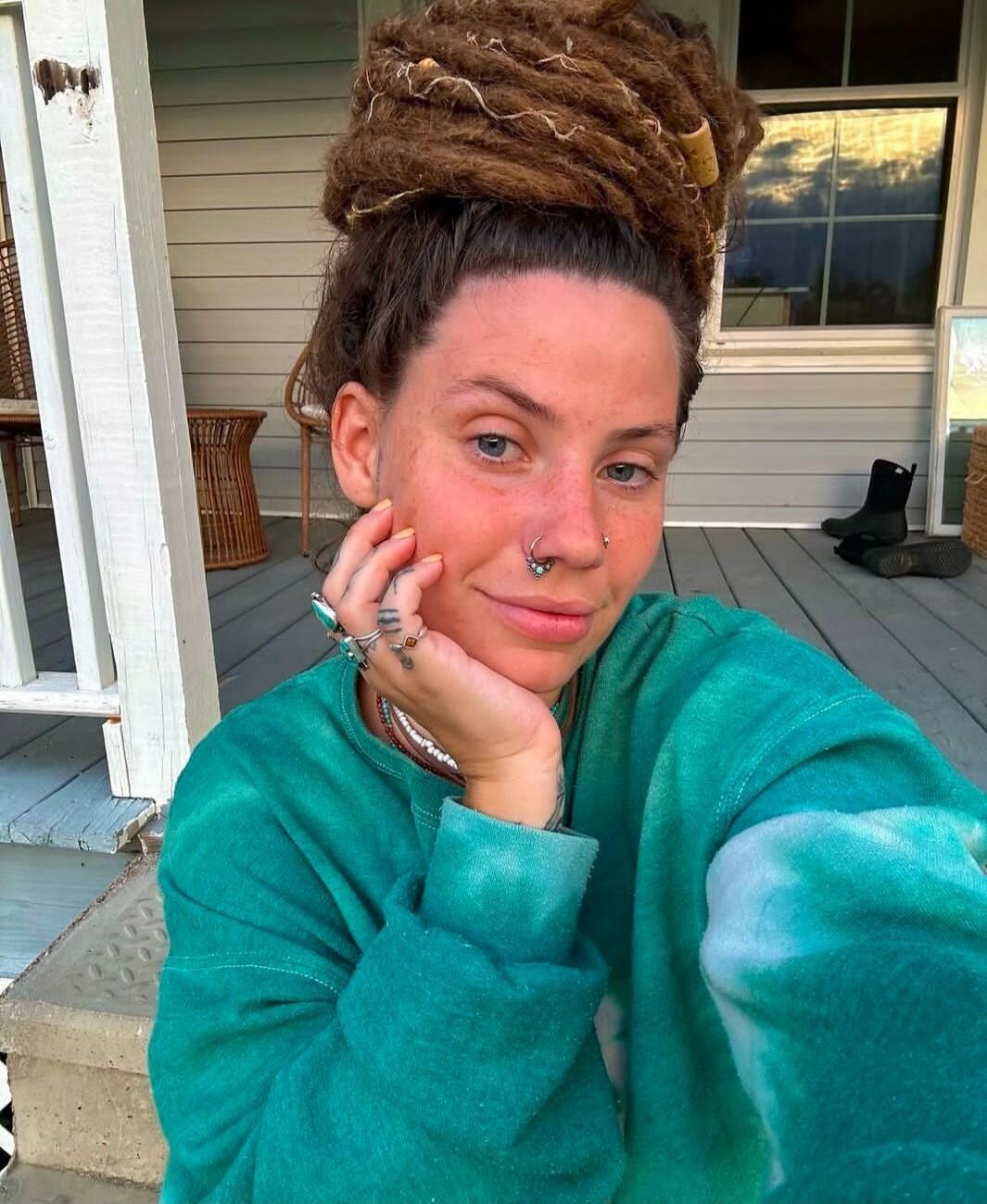 A woman with dreadlocks tied in a bun, wearing a green tie-dye sweatshirt, multiple rings, and nose piercings, is sitting on a porch, smiling slightly, with her hand resting on her chin.