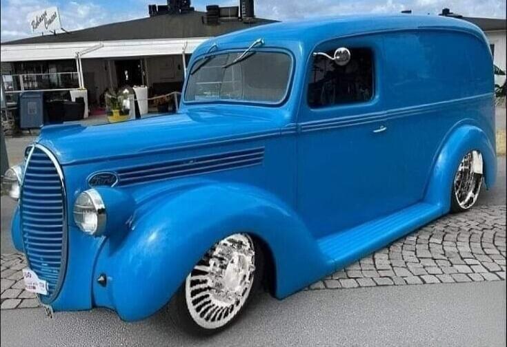 A bright blue vintage Ford panel van, possibly a custom hot rod or lowrider, is parked on a cobblestone street in front of a building with a sign that reads 'Bohuslän Kust'. The van features chrome wheels and a lowered suspension.