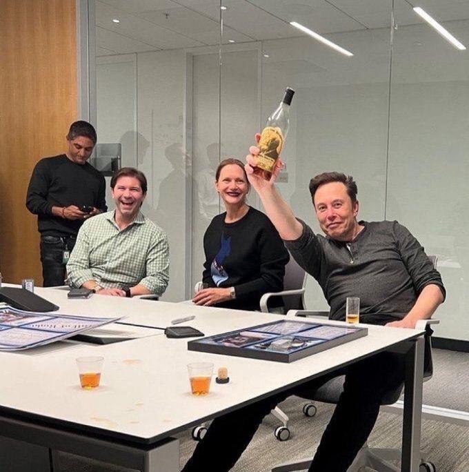 Elon Musk is seen holding up a bottle, celebrating with colleagues in an office setting.