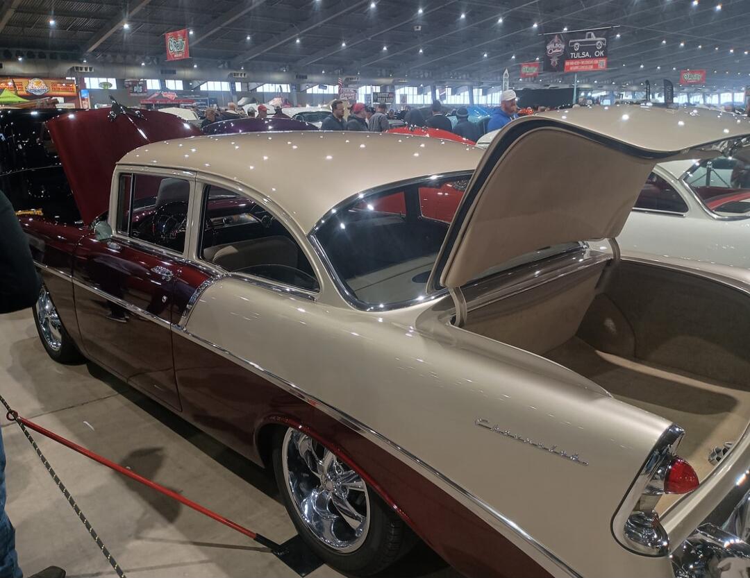 A classic two-tone Chevrolet car, possibly a Bel Air, at a car show. The hood and trunk are open, showcasing the vehicle. Other classic cars and people are visible in the background. Text on a banner reads 'TULSA, OK', and 'Chevrolet' is visible on the car's rear.