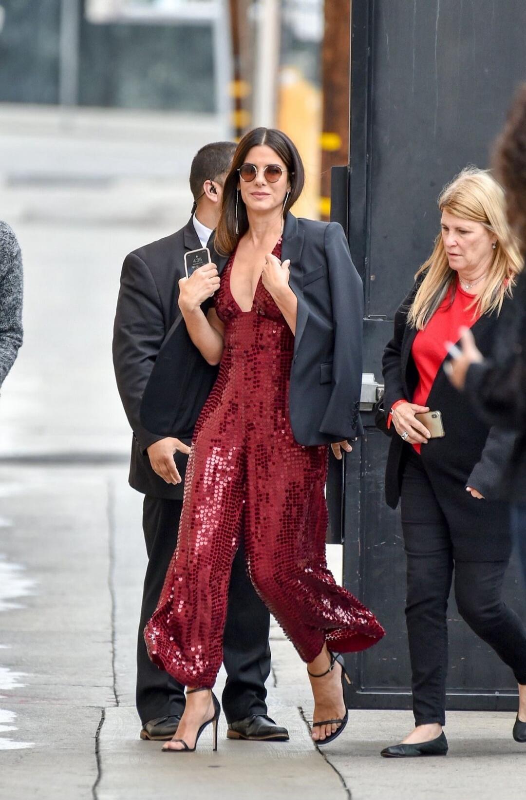 Woman in a red sequin jumpsuit with a black blazer, wearing sunglasses, walking on a city sidewalk. She is accompanied by others and appears to be photographed by several people.