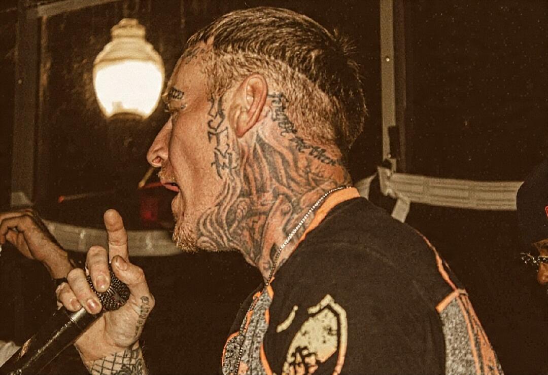 A man with numerous tattoos on his face and neck, holding a microphone and appearing to perform or speak. He has a cigarette in his mouth and is gesturing with his finger. The background is dark with a visible light fixture.
