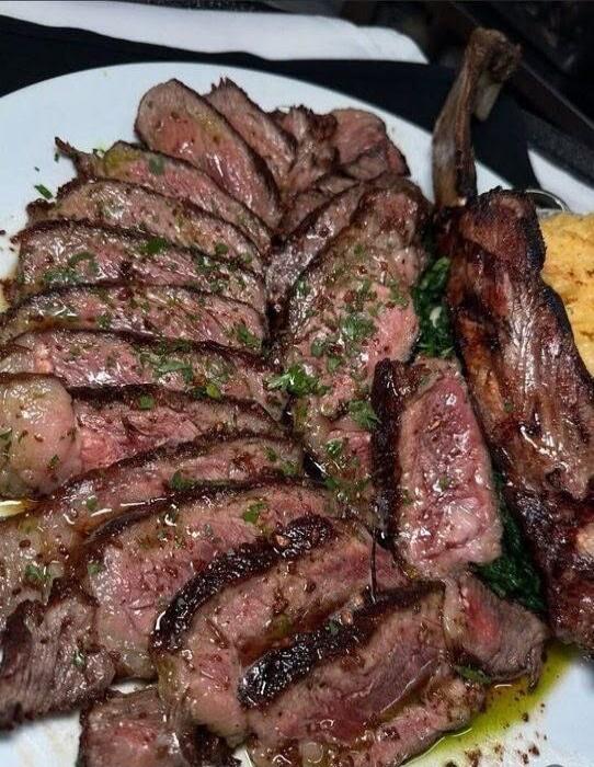 A close-up shot of a perfectly cooked and sliced steak, possibly a Tomahawk, served on a white plate. The meat is medium-rare, juicy, and garnished with fresh herbs. It is accompanied by some green vegetables and a yellow side dish, possibly mashed potatoes or polenta.
