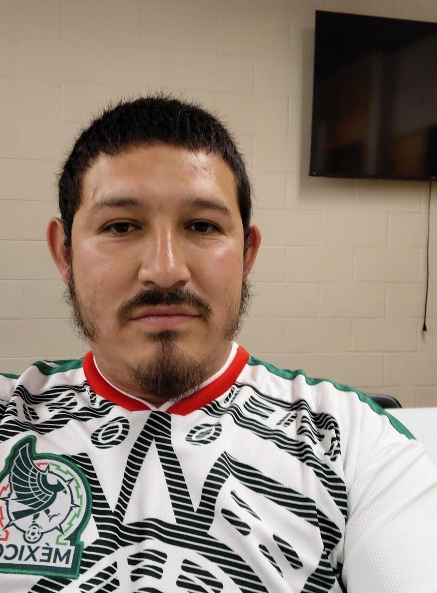 A man wearing a white, green, and red jersey with the word 'MEXICO' and an emblem on it. The background is a light-colored brick wall with a dark TV screen.