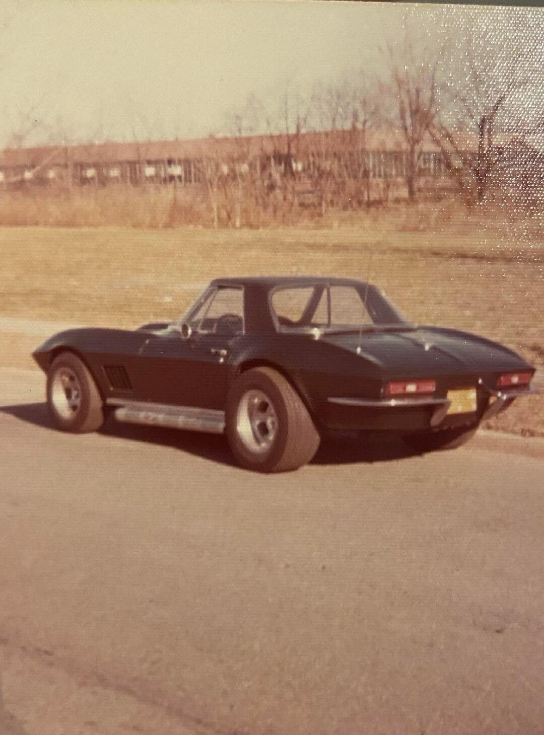 A black vintage sports car, likely a mid-1960s Corvette, parked on a road with an open field and trees in the background.