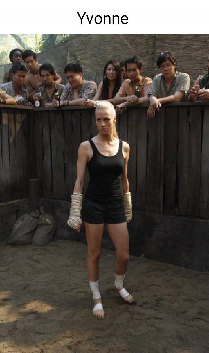 Yvonne. A woman in a fighting stance, with wrapped hands and feet, stands in a sandy pit. A crowd watches from above a wooden fence.