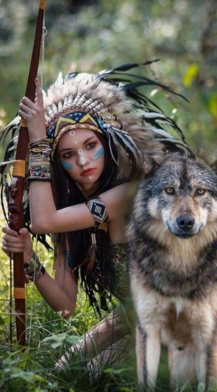 The image shows a young woman dressed as a Native American, wearing a feathered headdress and face paint. She is holding a bow and arrow. Beside her stands a wolf, looking directly at the camera. The scene is set in a forest.