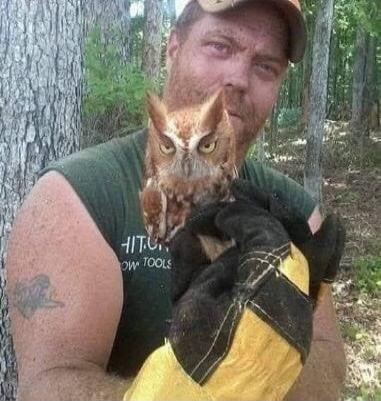 A man with a beard and a cap, covered in dirt, is holding a small owl with yellow eyes and prominent ear tufts. He is wearing work gloves. The text 'HIT.U. OW TOOLS' is visible on his green shirt. The background is a forest.