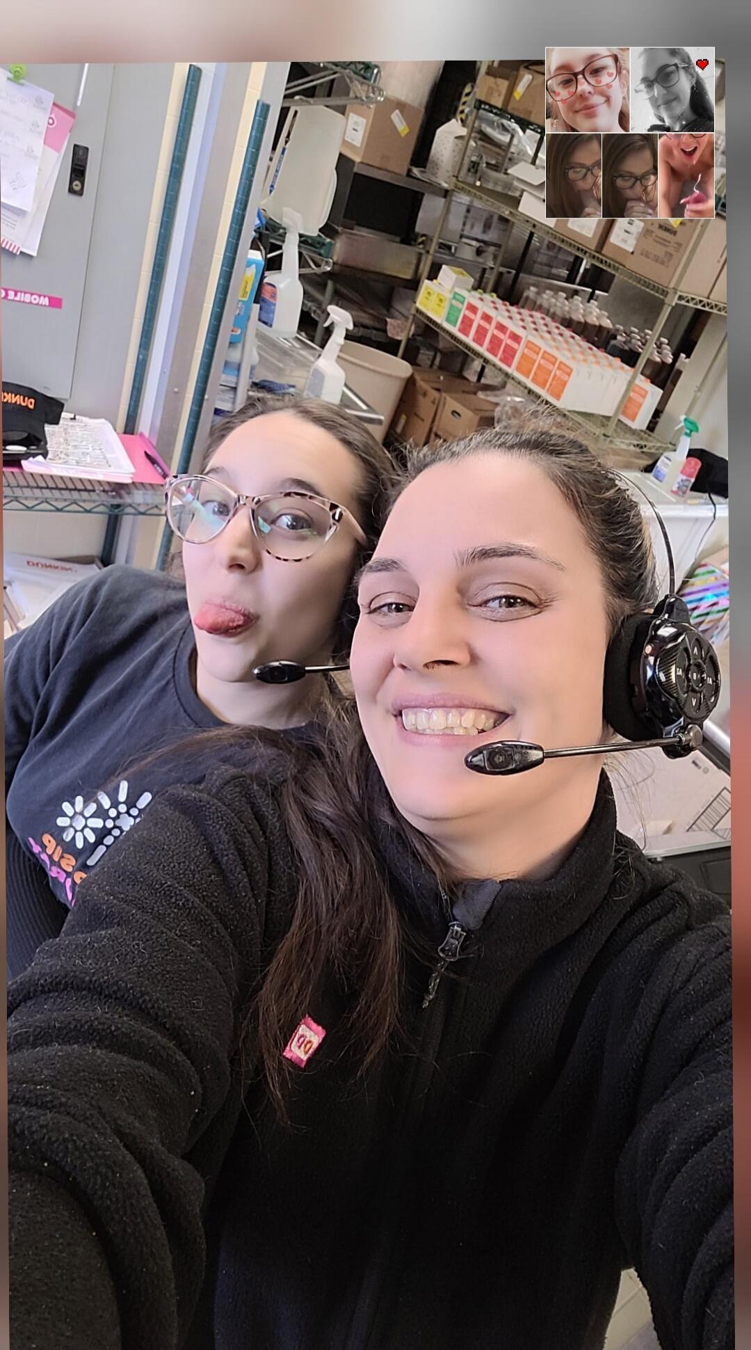 Two women wearing headsets take a selfie. One woman smiles at the camera, while the other makes a funny face by sticking out her tongue. In the background, shelves with various products are visible. A reversed 'DUNKIN' logo is visible on a cap, and a 'DD' logo is on one woman's jacket.