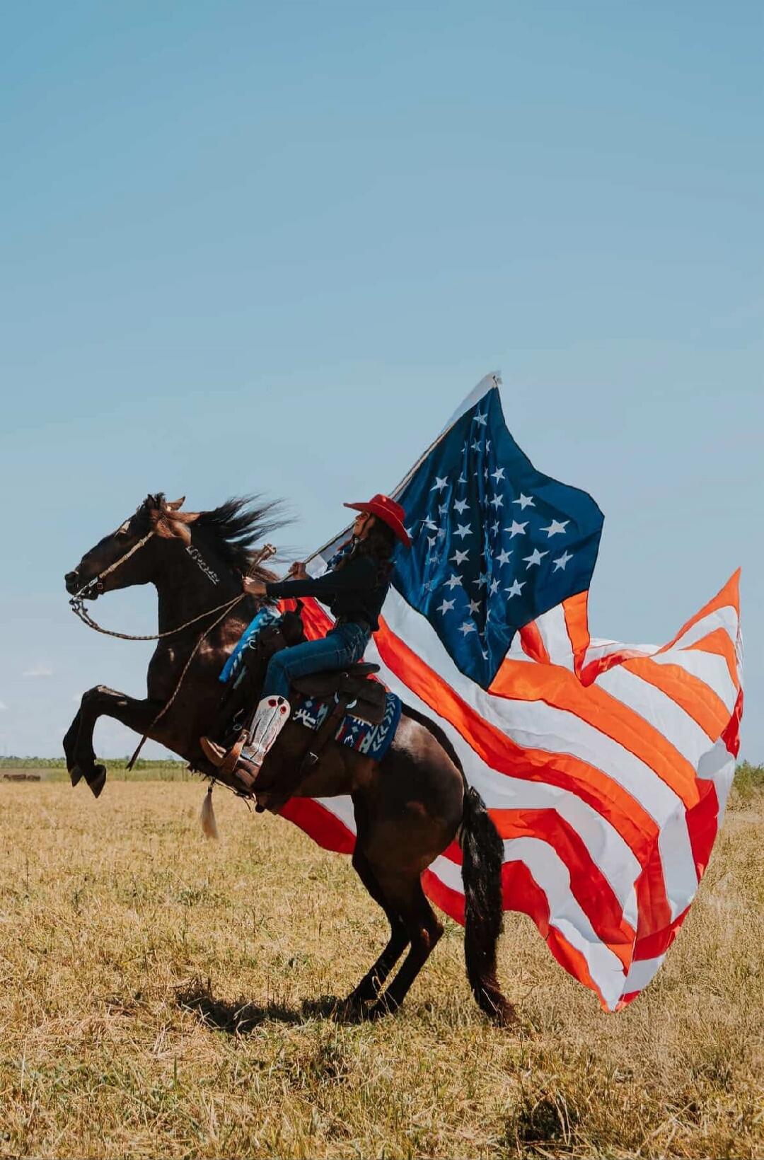 A person in a cowboy hat and attire riding a rearing horse, with a large American flag flowing behind them in an open field under a clear sky.