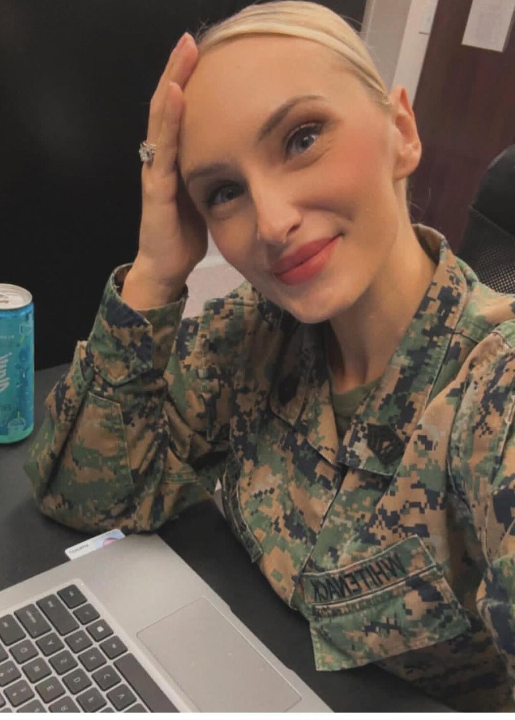 A blonde woman in a military uniform (camouflage pattern) is smiling at the camera. She is sitting at a desk with a laptop and a can. Her hand is resting on her forehead. A name tag with 'WHILEVCK' (reversed) is visible on her uniform.