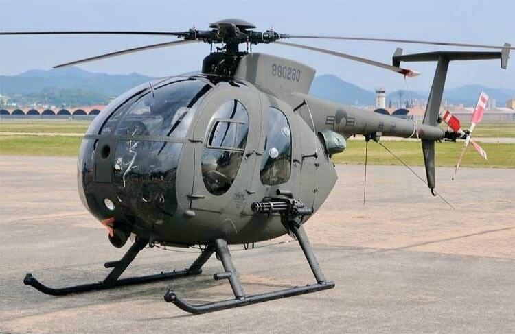 A military helicopter, likely an AH-6 Little Bird, is parked on an airfield. It is painted in a dark green or grey camouflage color and features a transparent bubble cockpit. The helicopter has a main rotor and a tail rotor, and is equipped with a minigun mounted on its side. The serial number '880280' is visible on the tail boom, along with a US m