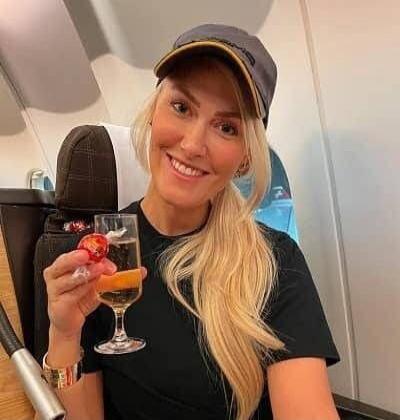 Photo of a smiling woman on an airplane holding a glass with an orange drink and a cherry/fruit garnish.