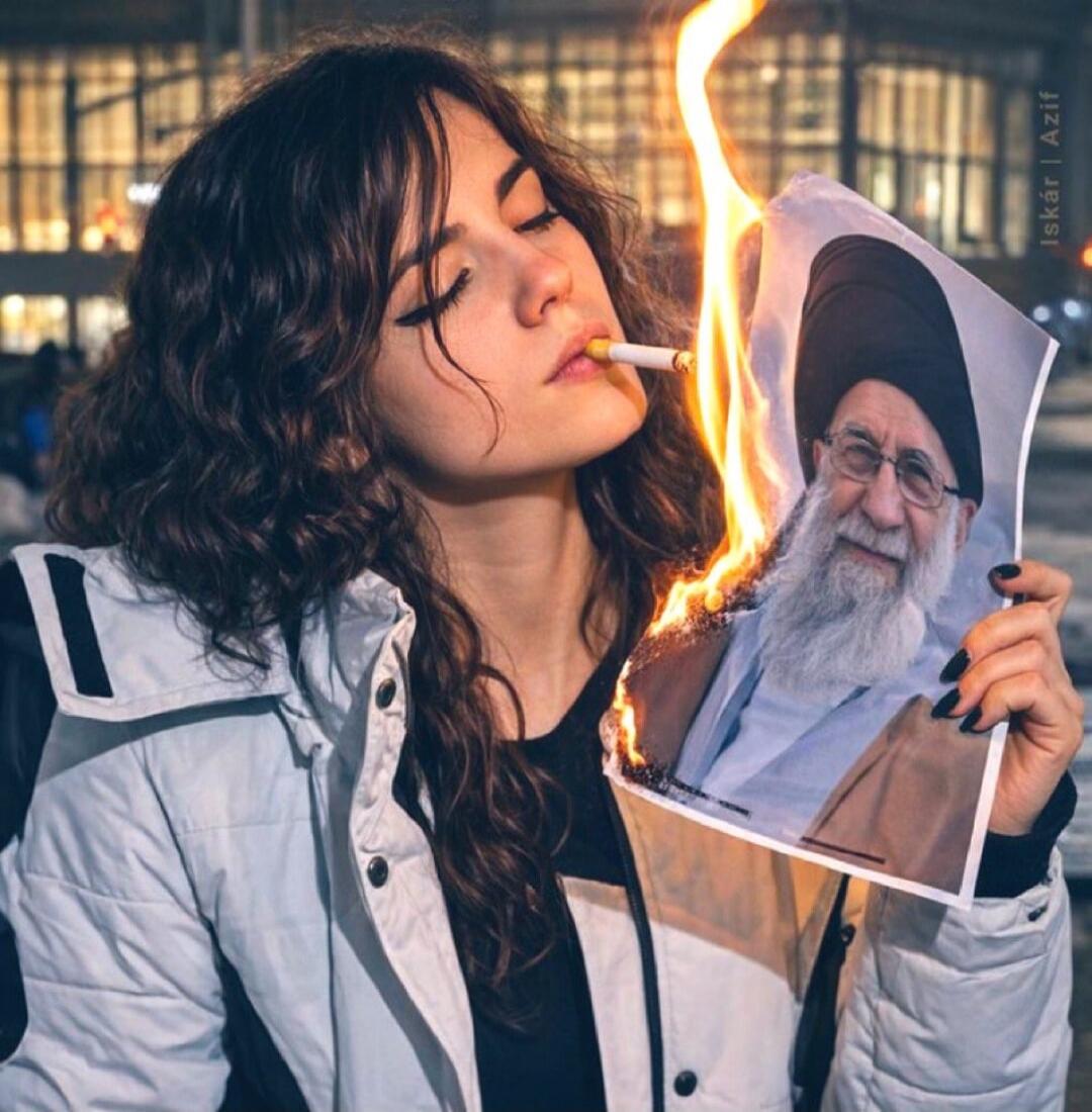 A young woman with a cigarette in her mouth is burning a printed photograph of a bearded man wearing a turban.