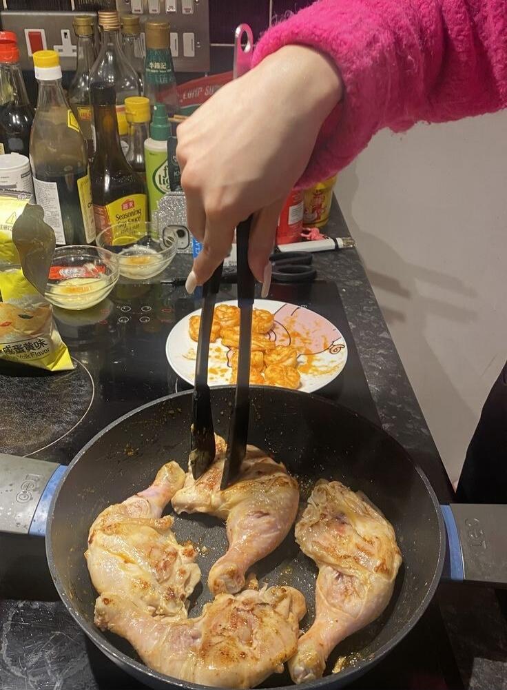 A person is cooking chicken drumsticks in a pan on a stovetop. The chicken is browning. In the background, there are various bottles of sauces and seasonings, including one labeled 'Seasoning Sauce'. A plate with cooked shrimp or similar items is also visible. A package with Chinese text '咸蛋黄味' (Salted Egg Yolk Flavor) is on the counter.
