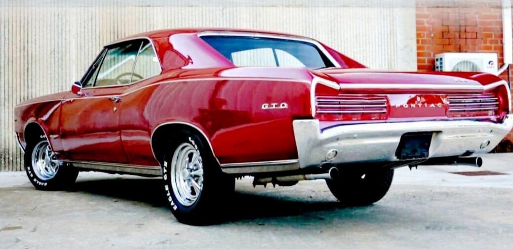 A classic red Pontiac GTO muscle car is parked, viewed from the rear three-quarter angle. The car features chrome bumpers and wheels, with 'G.T.O.' and 'PONTIAC' visible on the rear. The tires have 'GOOD YEAR EAGLE GT' written on them. The background shows a textured wall and a brick wall with an air conditioning unit.