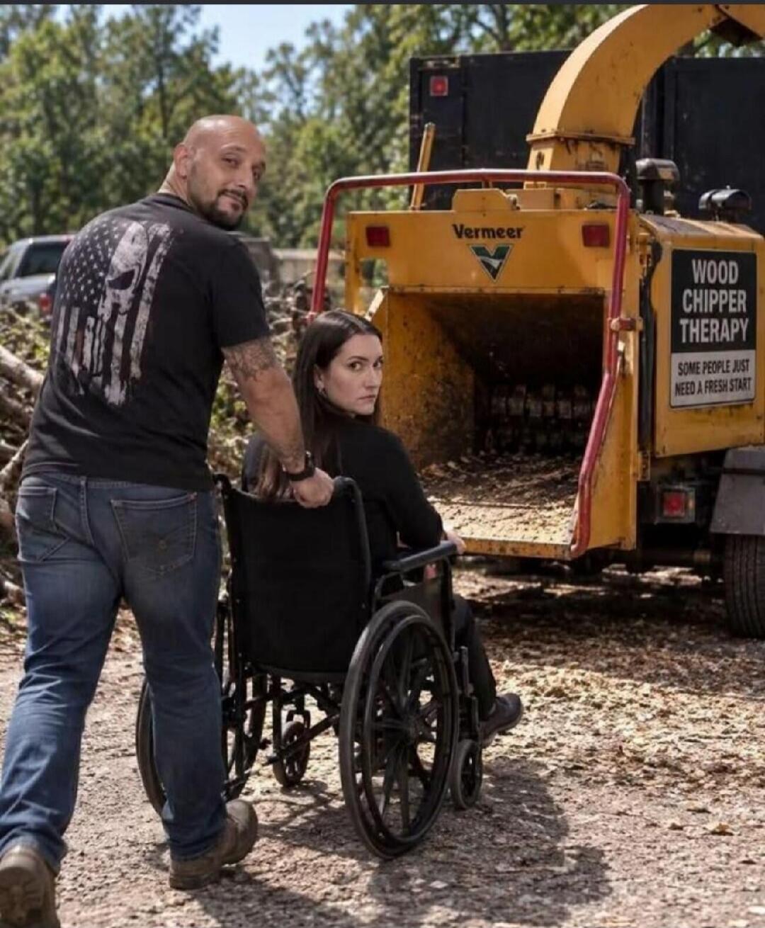 A man is pushing a woman in a wheelchair towards a yellow wood chipper. The woman is looking back at the camera with a serious expression, while the man is smiling. The wood chipper has text that reads 'Vermeer', 'WOOD CHIPPER THERAPY' and 'SOME PEOPLE JUST NEED A FRESH START'. The man is wearing a t-shirt with a Punisher skull and American flag de