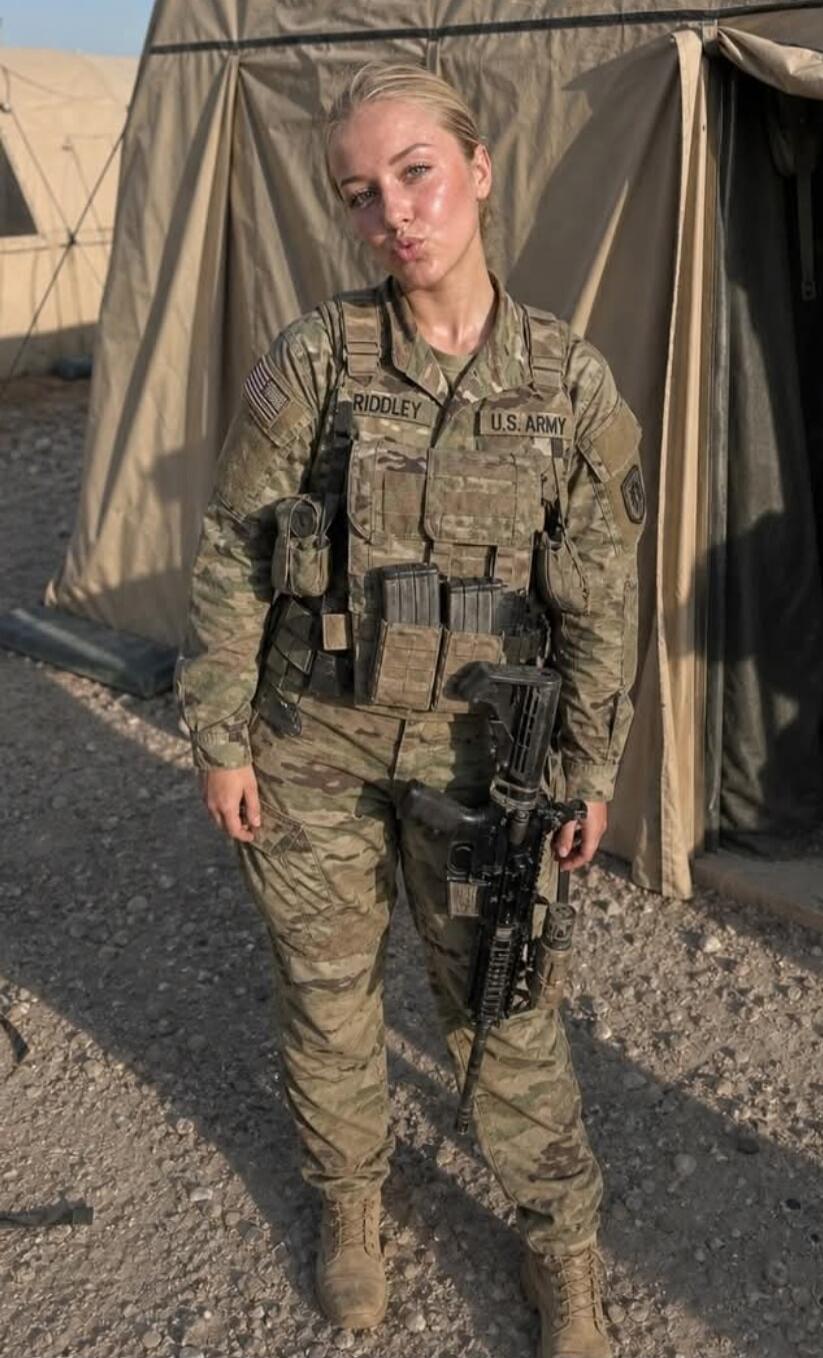 A young woman in a U.S. Army uniform, with the name 'RIDDLEY' and 'U.S. ARMY' visible on her uniform, makes a 'kiss face' expression while holding a rifle. She is standing outdoors in what appears to be a military camp.