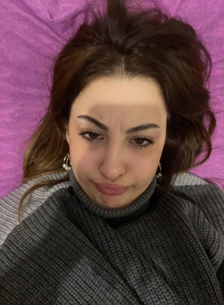 A young woman with dark hair and a grey turtleneck sweater is lying down, looking at the camera with a slightly pouting or displeased expression. She is wearing pearl earrings.