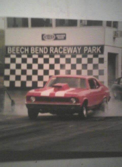 BEECH BEND RACEWAY PARK. A red muscle car with white racing stripes is doing a burnout on a drag strip, creating a cloud of smoke from its tires.