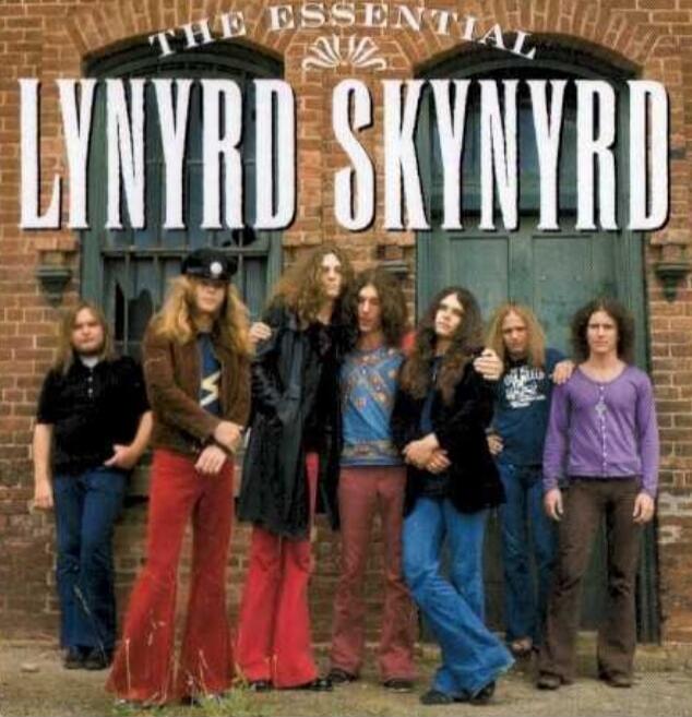 THE ESSENTIAL LYNYRD SKYNYRD. A group of seven men with long hair, dressed in 1970s style clothing, standing in front of a brick building with two doors.