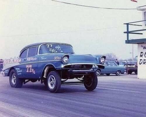 A blue vintage drag racing car, number 77 D/G, with '327ci' on the fender and 'California' on the side. Text on a building in the background reads 'ROD DRAG'.