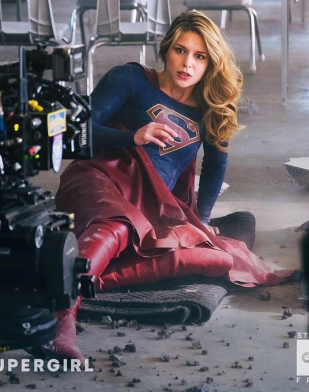 A woman dressed as Supergirl is on the ground, looking surprised or in distress, on what appears to be a film set. A camera rig is visible on the left. The text 'SUPERGIRL' is visible at the bottom left, and 'WINNER' on the camera rig.