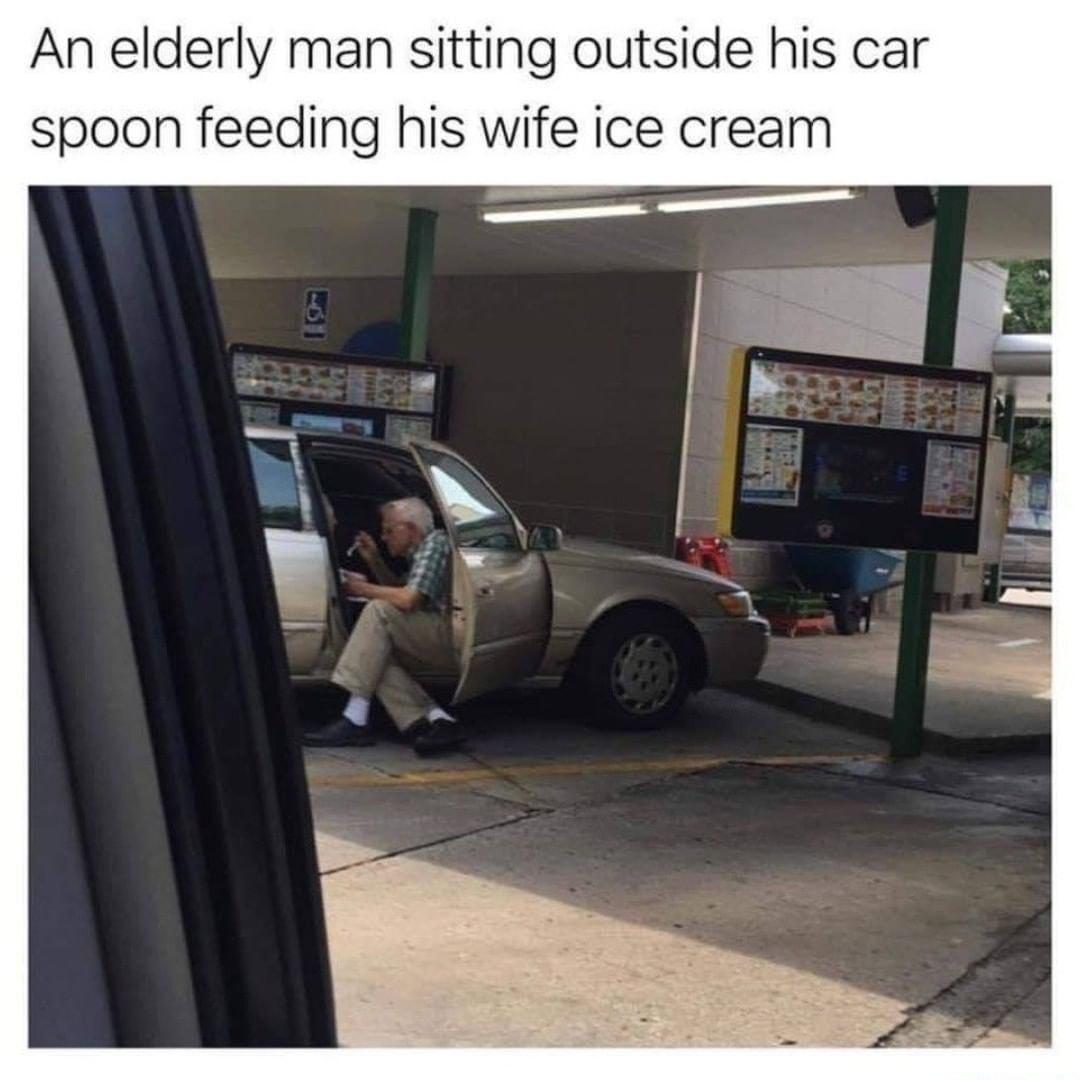 An elderly man sitting outside his car spoon feeding his wife ice cream