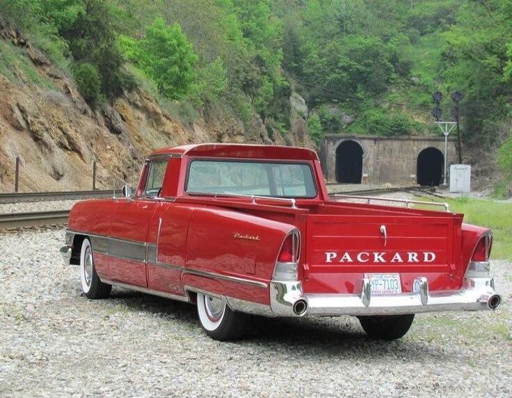 A red vintage Packard car, possibly a custom pickup conversion, is parked on gravel next to railroad tracks. The text 'PACKARD' is visible on the tailgate, and a license plate 'YP-7103' is also visible. In the background, there are green hills and two train tunnels.