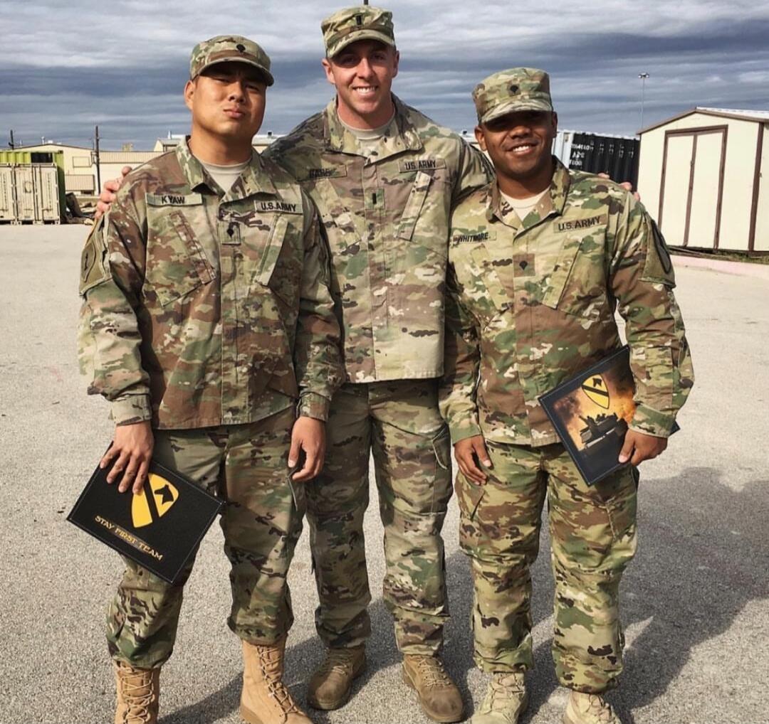 Three soldiers in camouflage uniforms posing outdoors, each holding a folder.