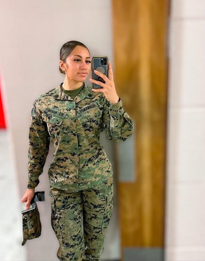 A young woman in a US Marine Corps digital camouflage uniform takes a mirror selfie with her phone. The name 'Hernandez' and 'PFC USMC' are visible on her uniform.