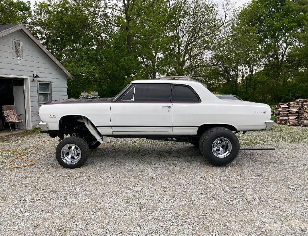A heavily modified white classic car, possibly a Chevrolet Malibu, with a lifted suspension and large wheels, featuring an exposed engine.