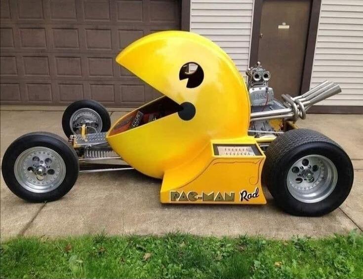A custom-built yellow hot rod designed to look like the video game character Pac-Man, with 'PAC-MAN Rod' written on its side. The car features a large, open-mouthed Pac-Man head as the body, exposed engine parts, and large rear wheels, parked in front of a garage.