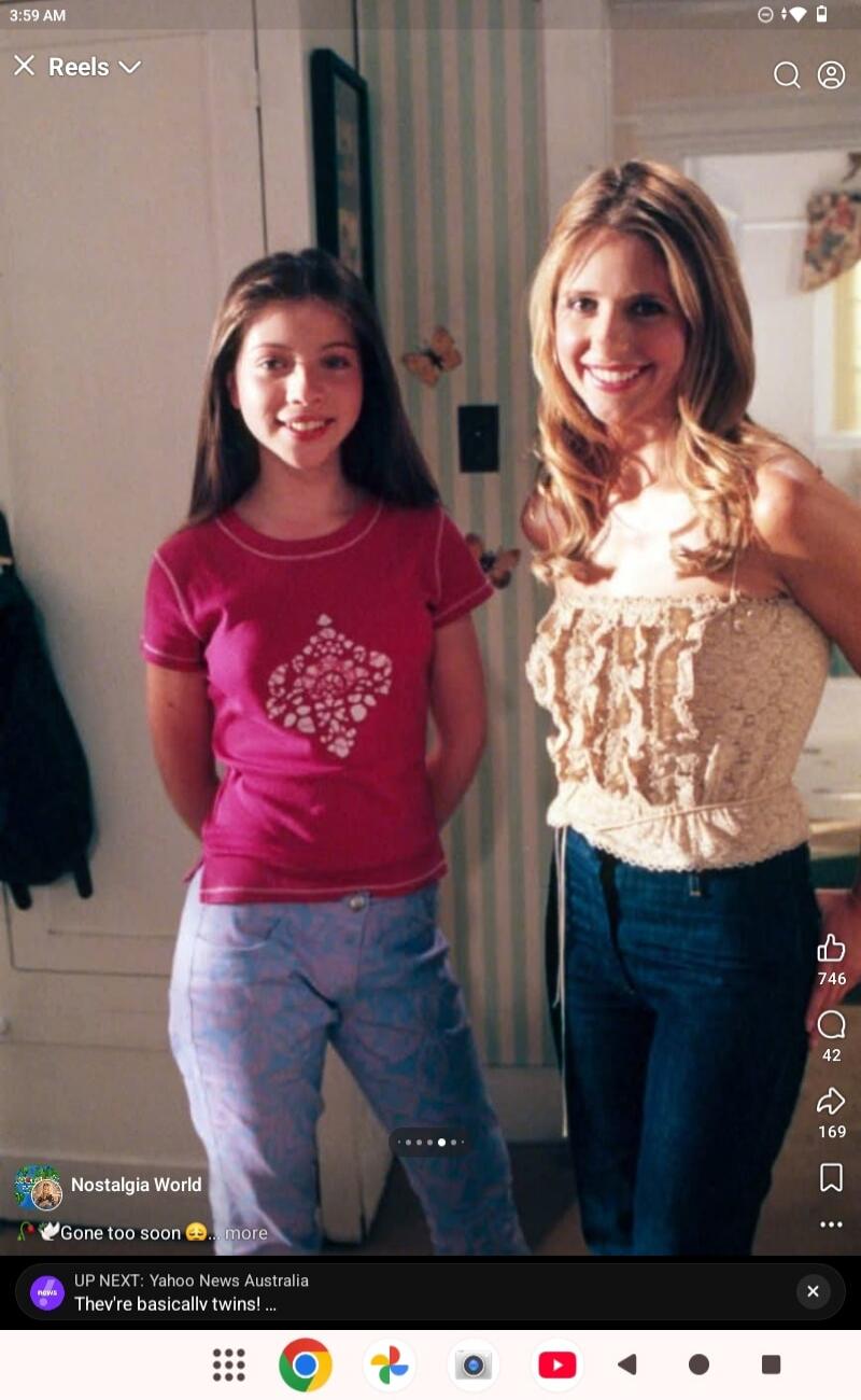 Two young women are standing in a room. The woman on the left is wearing a pink t-shirt and blue patterned pants. The woman on the right is wearing a strapless lace top and blue jeans. They are both smiling at the camera.