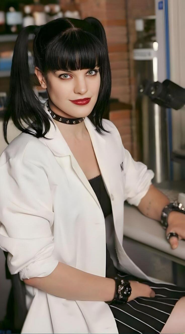 The image shows a woman with black hair styled in pigtails and bangs, wearing a white lab coat over a black top. She has striking green eyes and is wearing dark lipstick and a studded choker. Her left arm is resting on her lap, and she is wearing a large, dark watch with studs. Her right arm is bent, and she appears to be sitting in a chair, possib