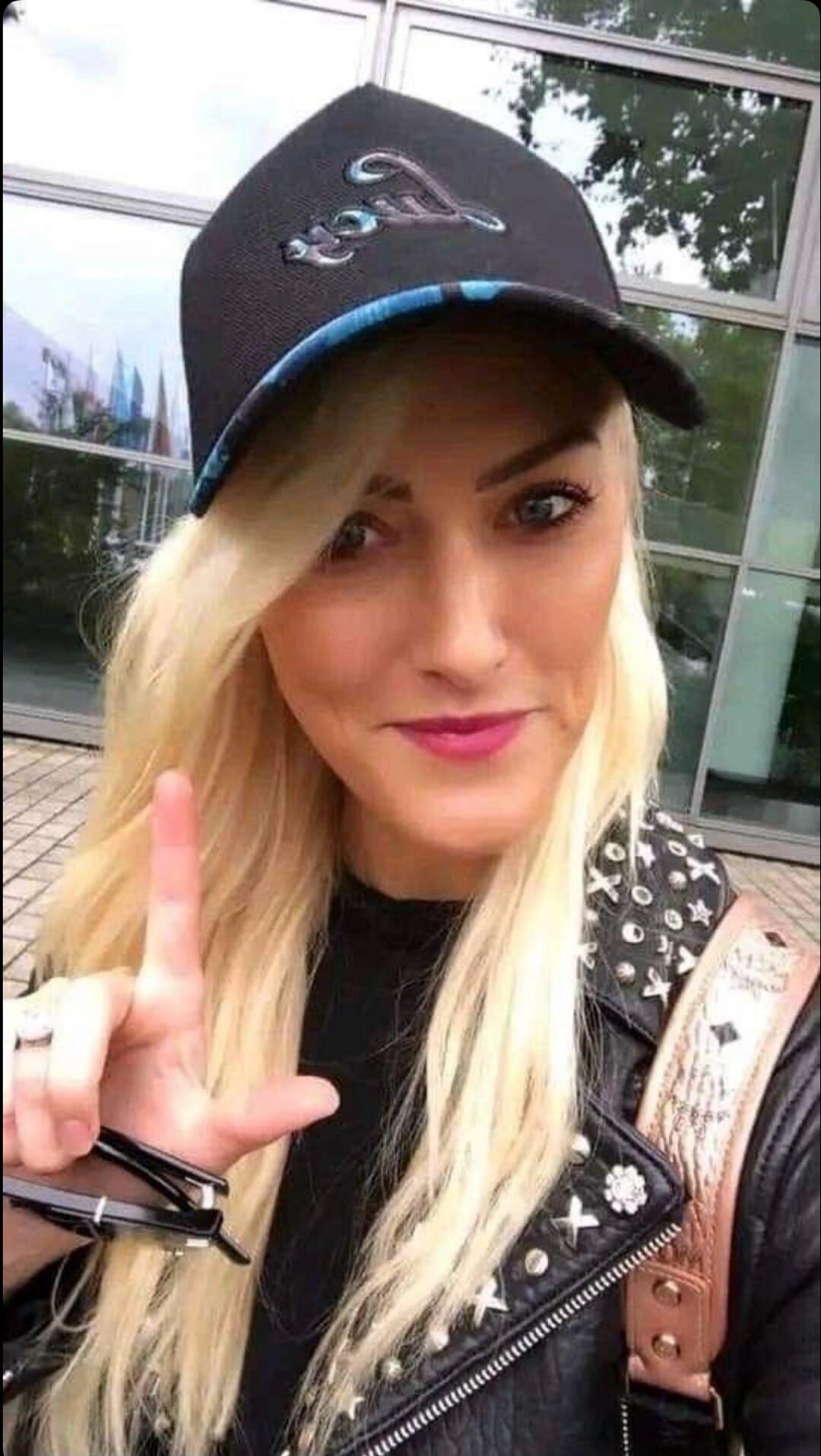 A blonde woman wearing a cap and a leather jacket, making an 'L' sign with her fingers.