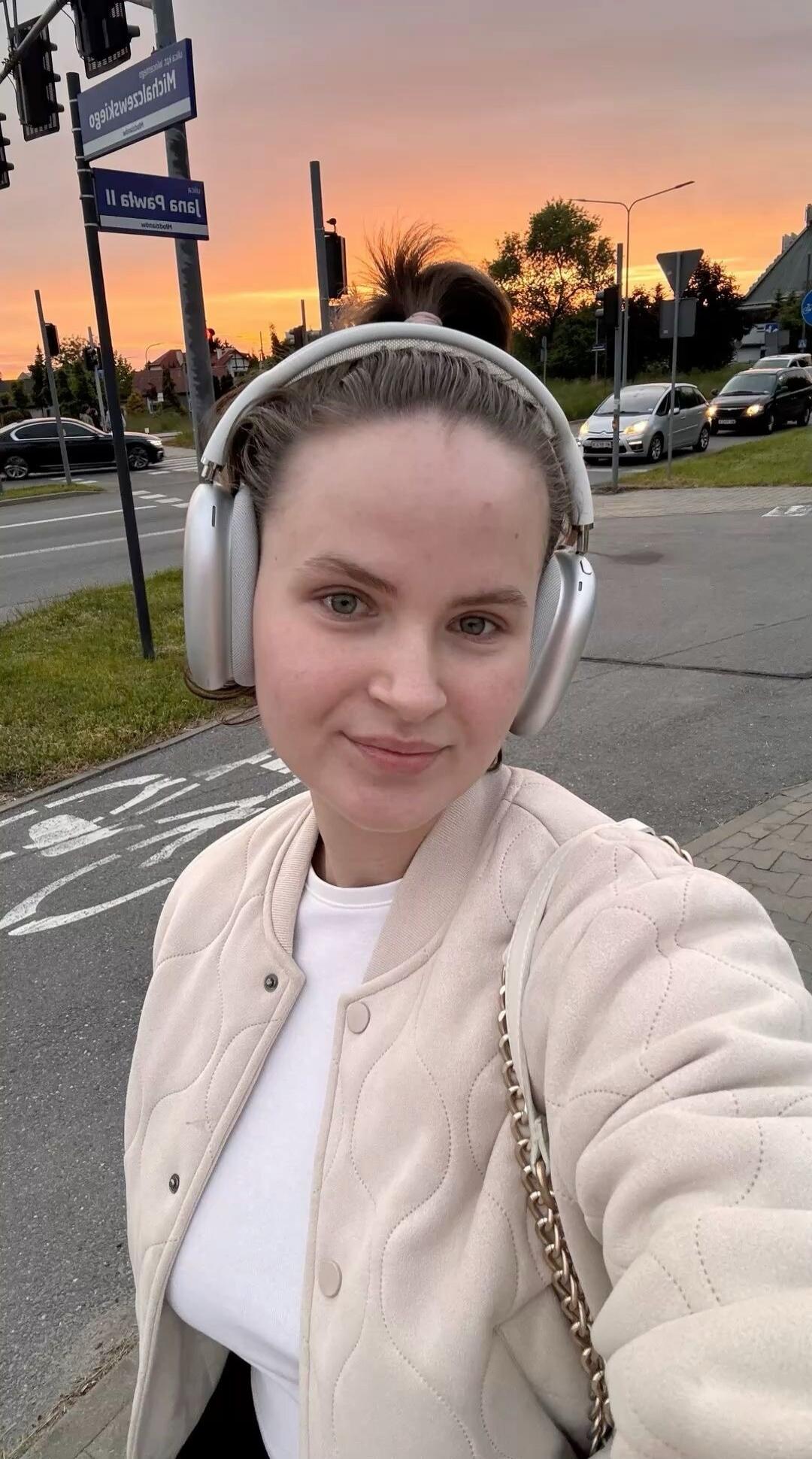 A young woman takes a selfie outdoors, wearing headphones and a light-colored quilted jacket. In the background, there are street signs with Polish names 'ul. Michała Piaszczyńskiego' and 'ul. Jana Pawła II', traffic lights, cars, and a bicycle lane marking on the asphalt. The sky shows the colors of a sunset or sunrise.