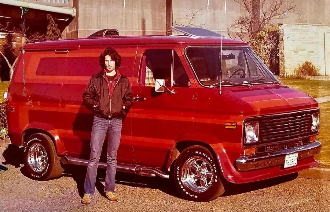 A man with long, dark hair stands next to a customized red van. The van features chrome wheels with 'RADIAL T/A' lettering, side exhaust pipes, and a sunroof. The license plate reads 'TH 3623'. The man is wearing a dark jacket, a red shirt, and jeans, with his hands in his pockets. The scene has a vintage aesthetic.
