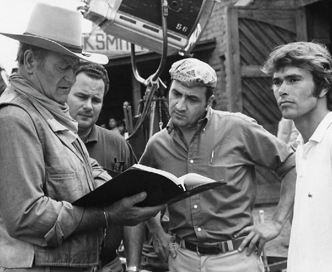 KSMIT 56. A group of men, including John Wayne, are on a film set. John Wayne, wearing a cowboy hat and a scarf, is holding and reading from a book or script. Other crew members are looking on, one with a patterned cap, another in a white polo shirt. A large film camera is visible in the background.