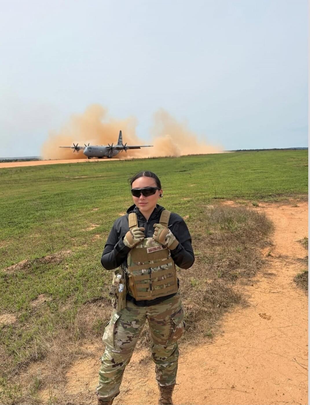 A woman in tactical gear stands in a field with a military transport plane kicking up dust in the background.