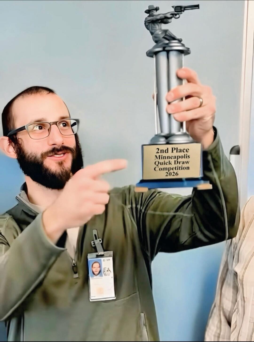 A man with glasses holding up a trophy that says '2nd Place Minneapolis Quick Draw Competition 2026'. He is pointing at it. He also wears an ID badge with a photo.