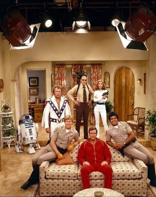 A group of iconic 1970s and early 1980s TV and movie characters, including R2-D2, Captain America, officers from CHiPs, Steve Austin from The Six Million Dollar Man, and other action/detective figures, are gathered on a studio set with large overhead lights.