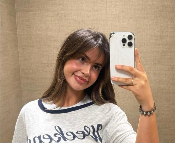 A young woman taking a mirror selfie wearing a white T-shirt with dark trim and a bracelet, smiling at the camera.