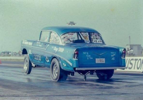 A blue vintage drag racing car, possibly a 1955-57 Chevrolet, is captured on a drag strip. The car features custom paint and various racing decals on its body and rear window. Visible text includes 'Shop SHOP' on the side and 'Built by Don & Tim' on the rear. A partial sign 'VISTON' (likely part of a track name like 'HOUSTON') is visible in the bac