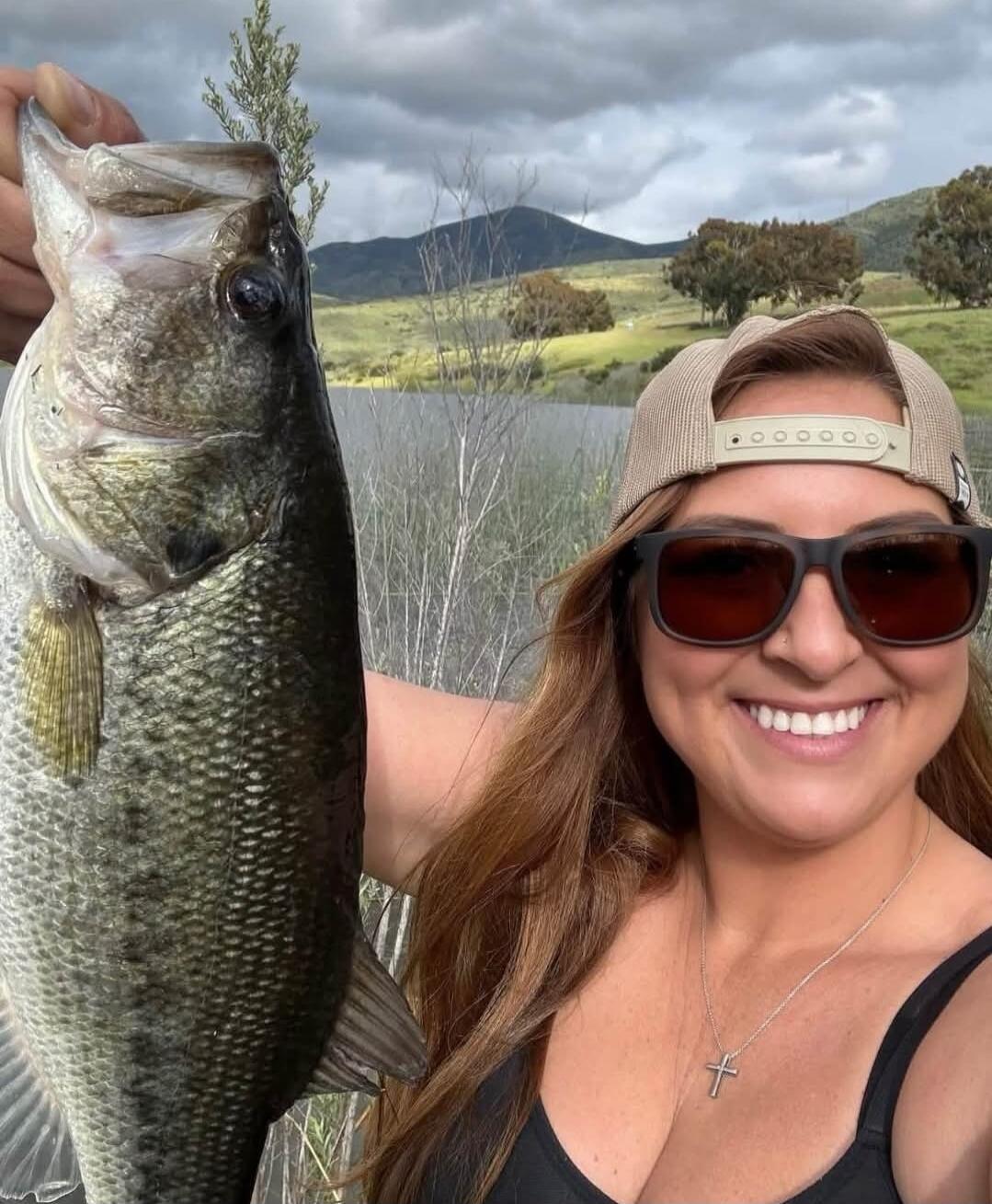 A woman smiling and holding a large bass fish. She is wearing sunglasses and a cap. In the background, there is a lake and mountains under a cloudy sky.