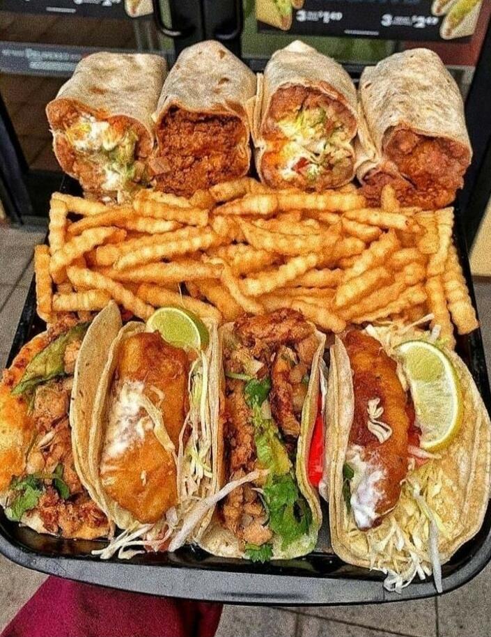 A tray filled with various Mexican-inspired fast food items, including four large burritos, a generous portion of golden french fries, and four tacos with different fillings such as fried fish and seasoned meat, garnished with lime wedges.