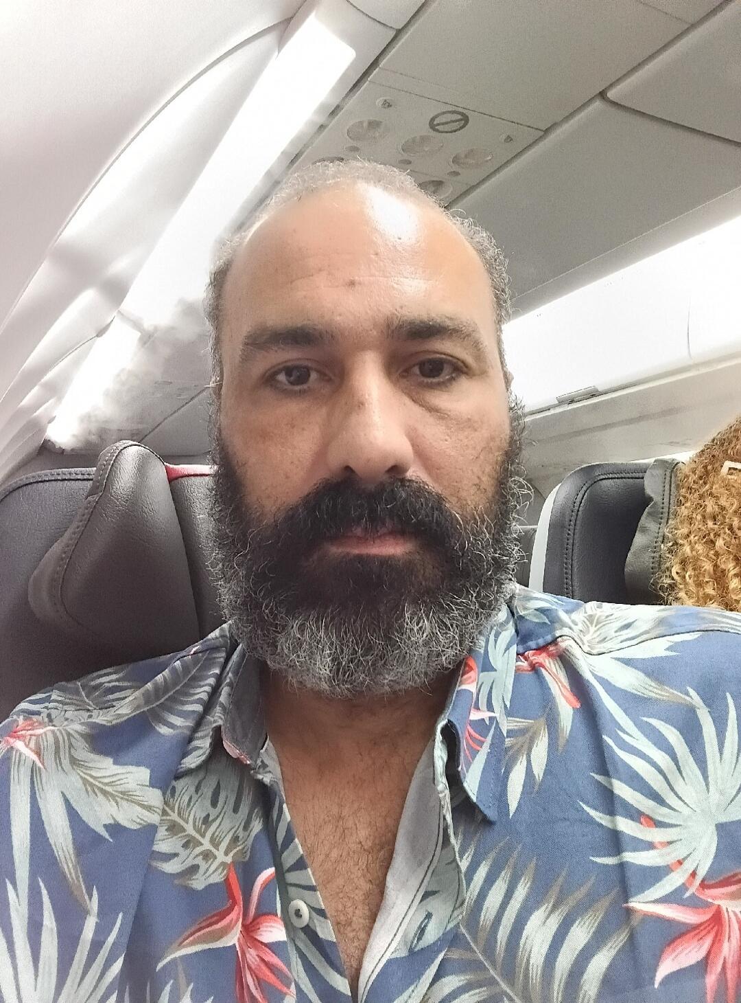 A man with a beard is taking a selfie on an airplane. He is wearing a blue Hawaiian-style shirt with red and green leaf patterns. There is some condensation or fog visible in the cabin near the window. Another person's curly hair is visible in the adjacent seat.
