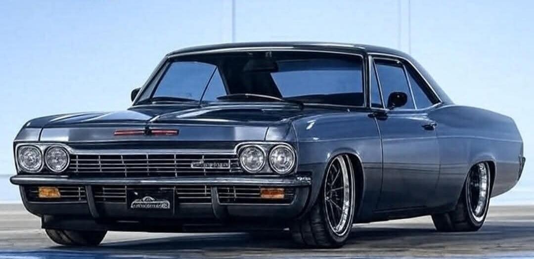 A classic dark grey or black Chevrolet Impala, possibly from the mid-1960s, is shown from the front-left angle. The car has a lowered suspension, custom wheels with chrome lips, and a sleek, polished appearance. The background is a plain light blue, suggesting a studio or outdoor setting with a clear sky.