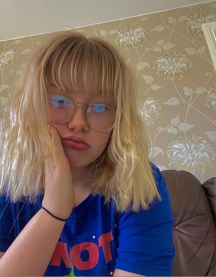 A young person with blonde hair and glasses, pouting and resting their chin on their hand. Text on t-shirt: 'HOT' (partially visible, reversed).
