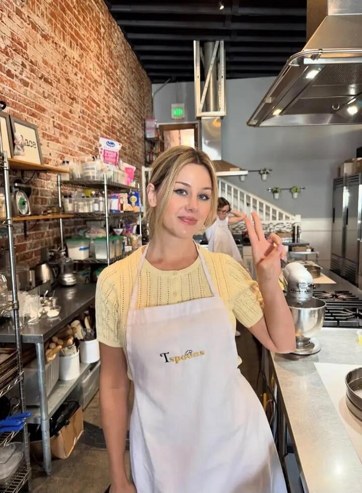 A young woman in a yellow top and a white apron with 'Tspoons' embroidered on it, making a peace sign in a kitchen. On a framed picture on the shelf, the word 'Lace' is visible.
